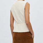 A woman shown from the back wearing a sleeveless cream peplum top with a matching tie belt and a brown skirt, set against a plain light gray studio background.
