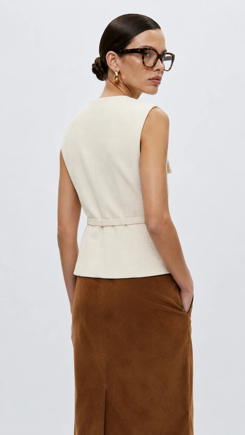 A woman shown from the back wearing a sleeveless cream peplum top with a matching tie belt and a brown skirt, set against a plain light gray studio background.