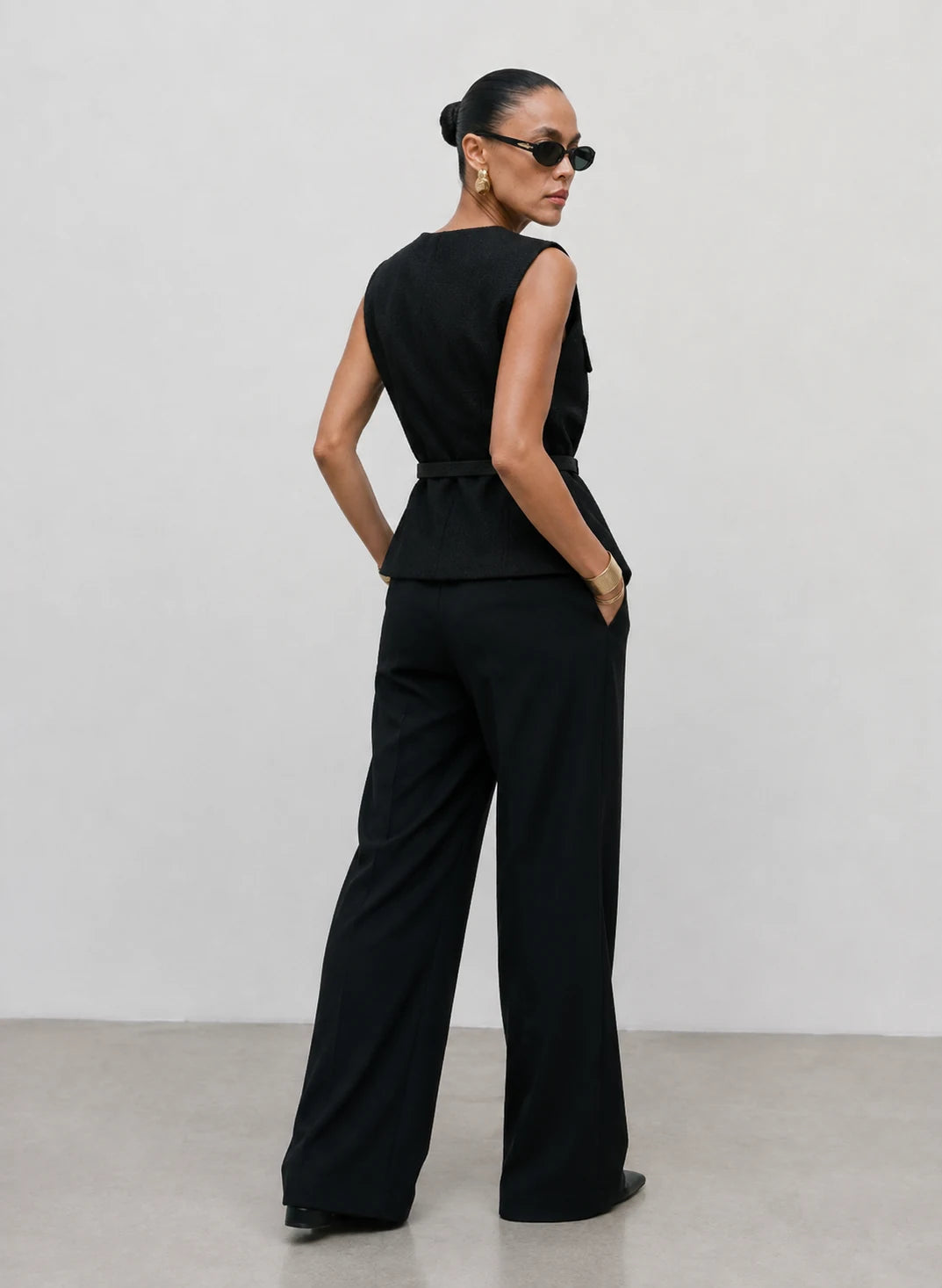 A model wearing a black sleeveless jumpsuit with a defined waist and wide-leg trousers, photographed from the back against a minimalist light gray studio background.