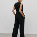 A model wearing a black sleeveless jumpsuit with a defined waist and wide-leg trousers, photographed from the back against a minimalist light gray studio background.