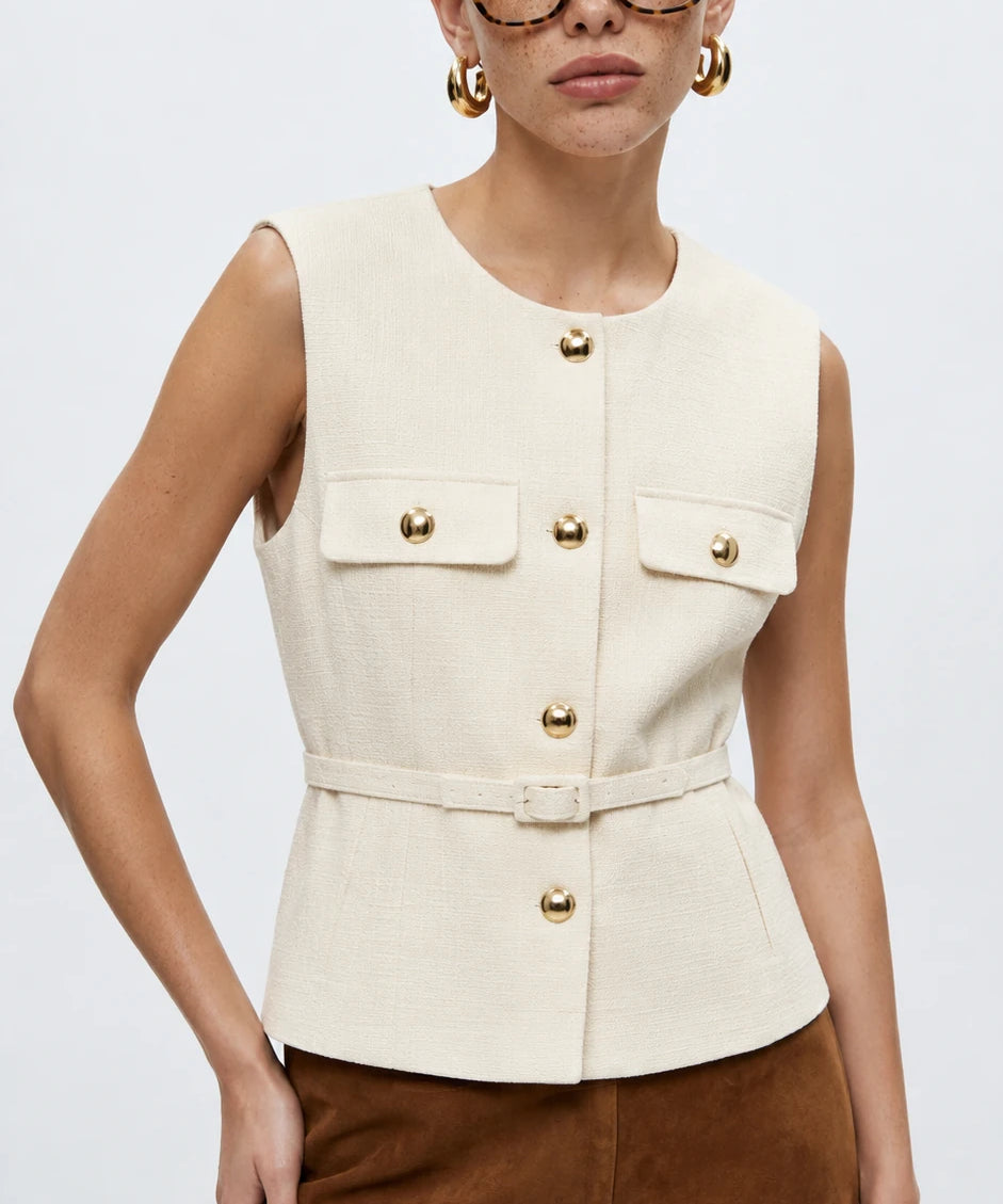 A woman modeling a sleeveless cream-colored tailored vest with gold-tone buttons and flap pockets, paired with brown trousers, against a plain light gray studio background.