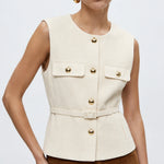 A woman modeling a sleeveless cream-colored tailored vest with gold-tone buttons and flap pockets, paired with brown trousers, against a plain light gray studio background.