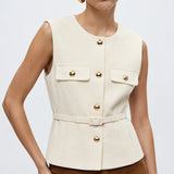 A woman modeling a sleeveless cream-colored tailored vest with gold-tone buttons and flap pockets, paired with brown trousers, against a plain light gray studio background.