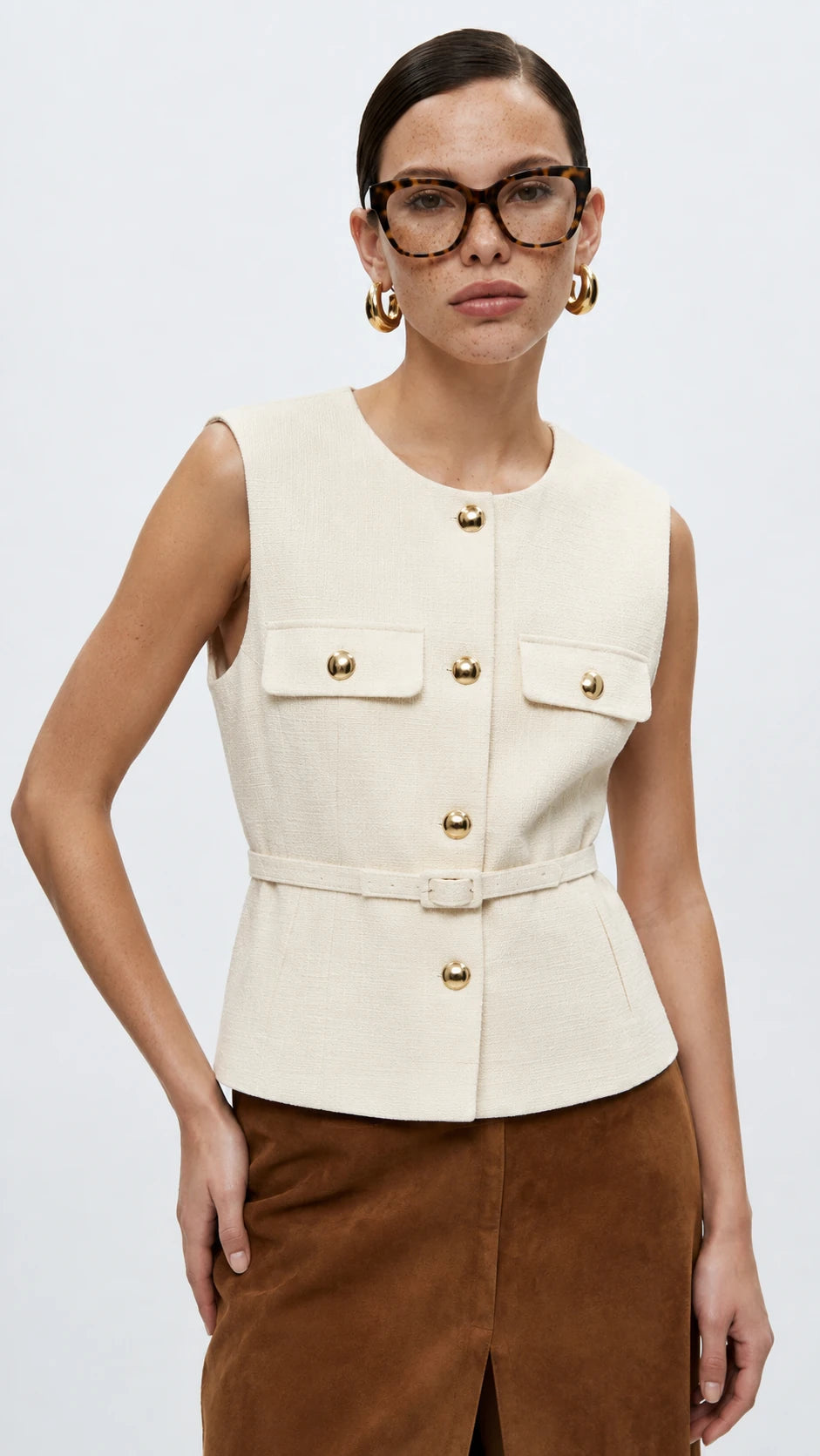 A woman modeling a sleeveless cream-colored tailored vest with gold-tone buttons and flap pockets, paired with brown trousers, against a plain light gray studio background.