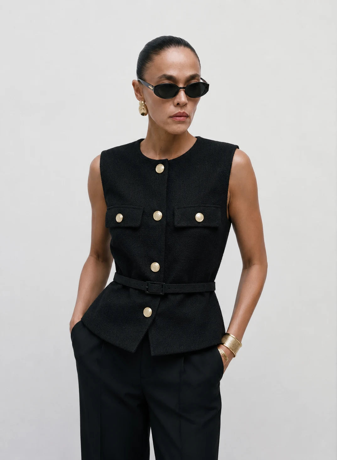 A model stands against a light gray studio background wearing a dark navy double-breasted blazer with gold buttons, wide-leg gray trousers, black sunglasses, and carrying a black handbag.