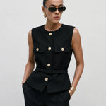 A model stands against a light gray studio background wearing a dark navy double-breasted blazer with gold buttons, wide-leg gray trousers, black sunglasses, and carrying a black handbag.
