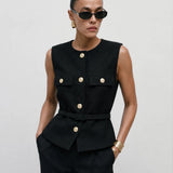 A model stands against a light gray studio background wearing a dark navy double-breasted blazer with gold buttons, wide-leg gray trousers, black sunglasses, and carrying a black handbag.