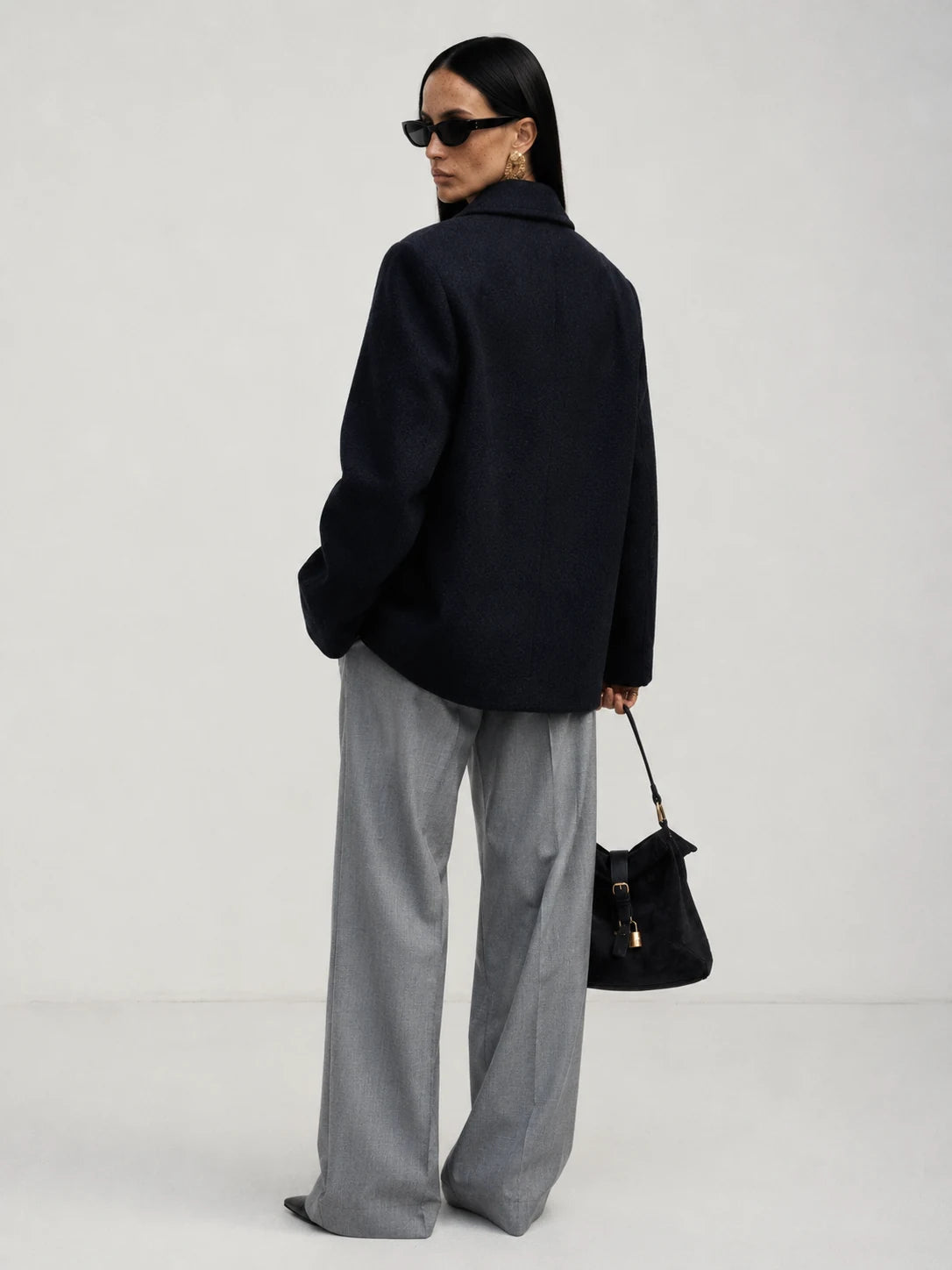 A woman is shown from behind wearing an oversized brown knit sweater, wide-leg light blue jeans, and holding a dark brown leather handbag against a plain light gray studio background.