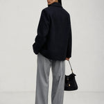 A woman is shown from behind wearing an oversized brown knit sweater, wide-leg light blue jeans, and holding a dark brown leather handbag against a plain light gray studio background.