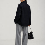 A woman is shown from behind wearing an oversized brown knit sweater, wide-leg light blue jeans, and holding a dark brown leather handbag against a plain light gray studio background.
