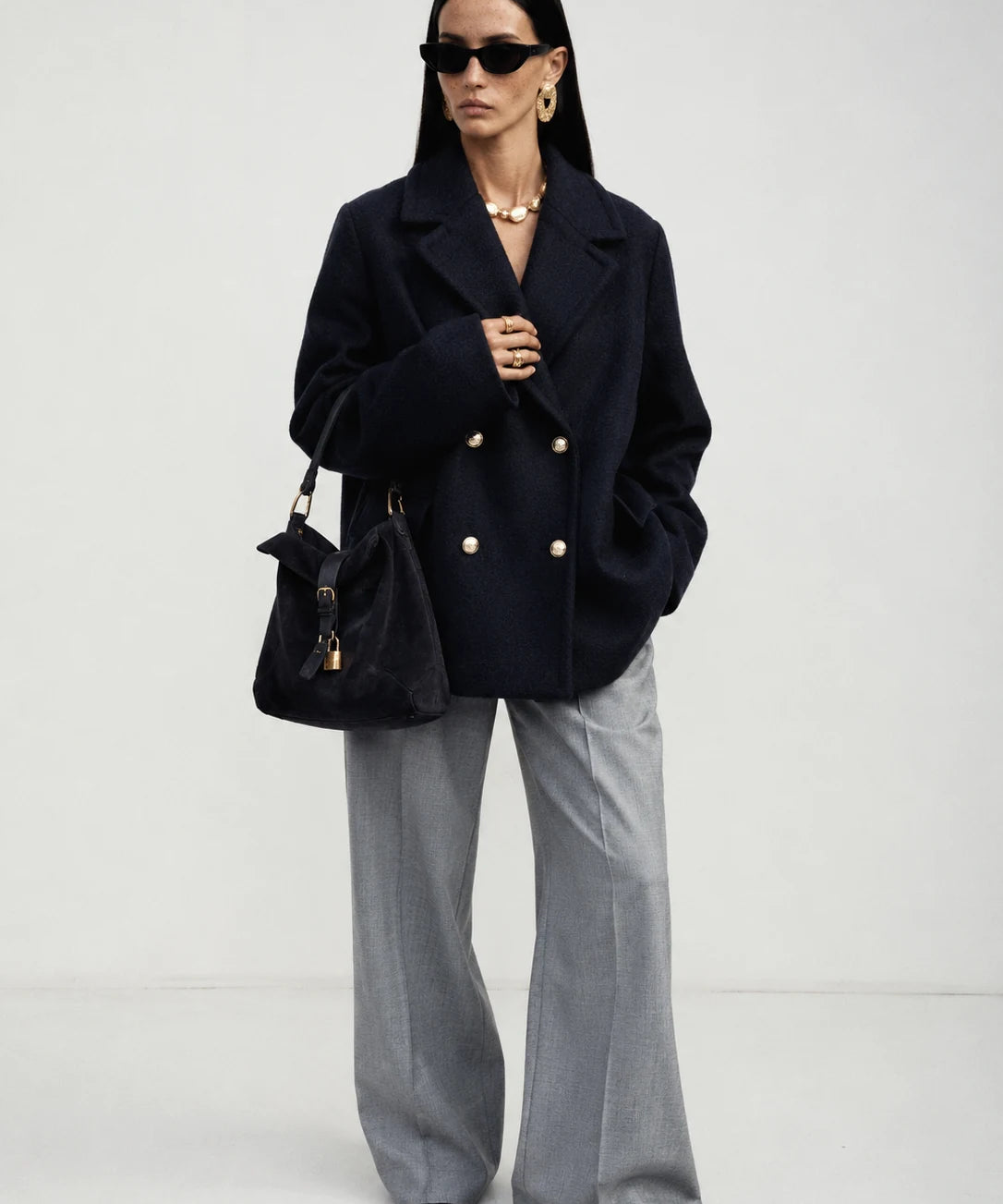 A model stands against a light gray studio background wearing a dark navy double-breasted blazer with gold buttons, wide-leg gray trousers, black sunglasses, and carrying a black handbag.