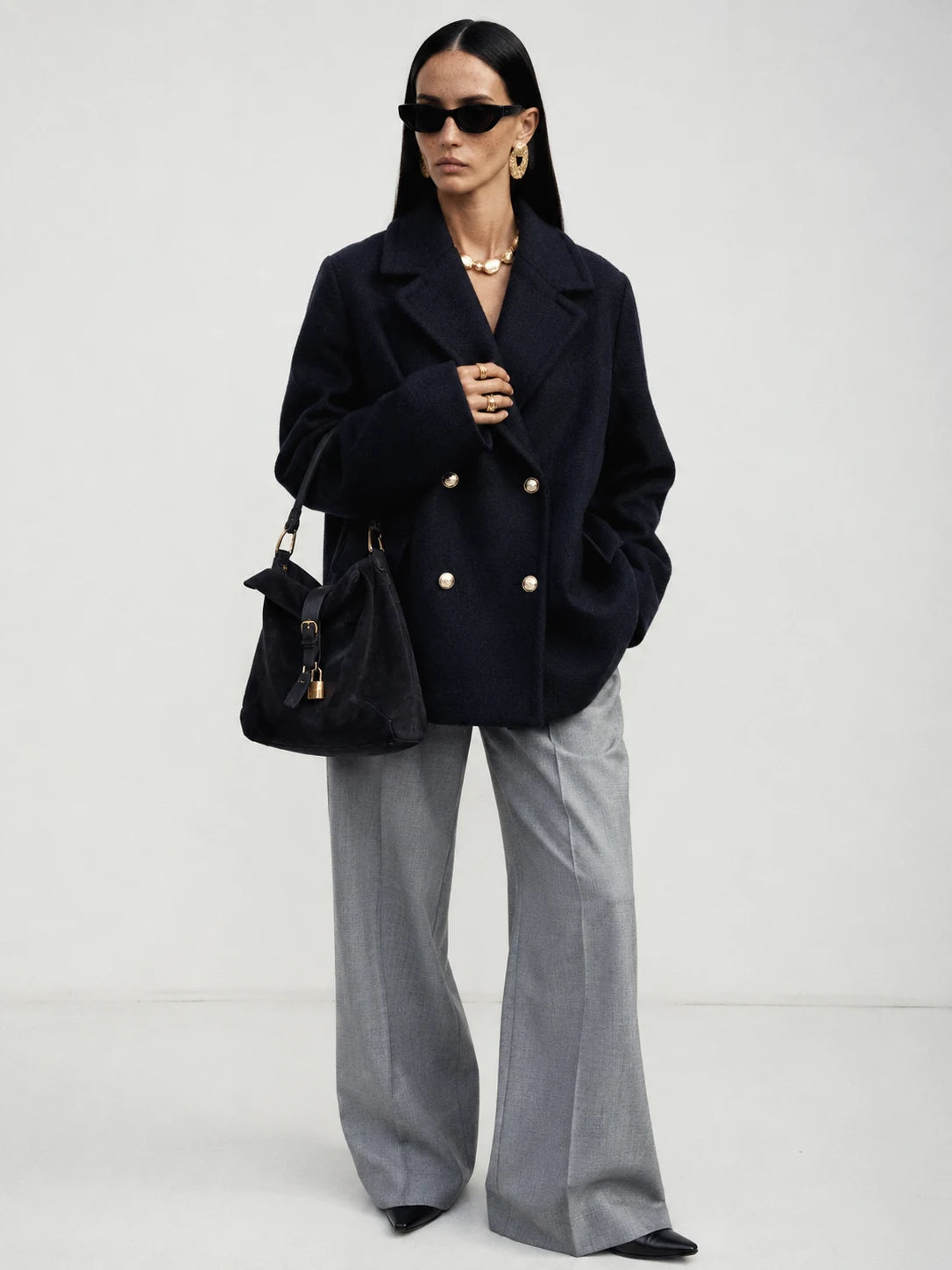 A model stands against a light gray studio background wearing a dark navy double-breasted blazer with gold buttons, wide-leg gray trousers, black sunglasses, and carrying a black handbag.