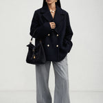 A model stands against a light gray studio background wearing a dark navy double-breasted blazer with gold buttons, wide-leg gray trousers, black sunglasses, and carrying a black handbag.