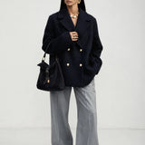 A model stands against a light gray studio background wearing a dark navy double-breasted blazer with gold buttons, wide-leg gray trousers, black sunglasses, and carrying a black handbag.