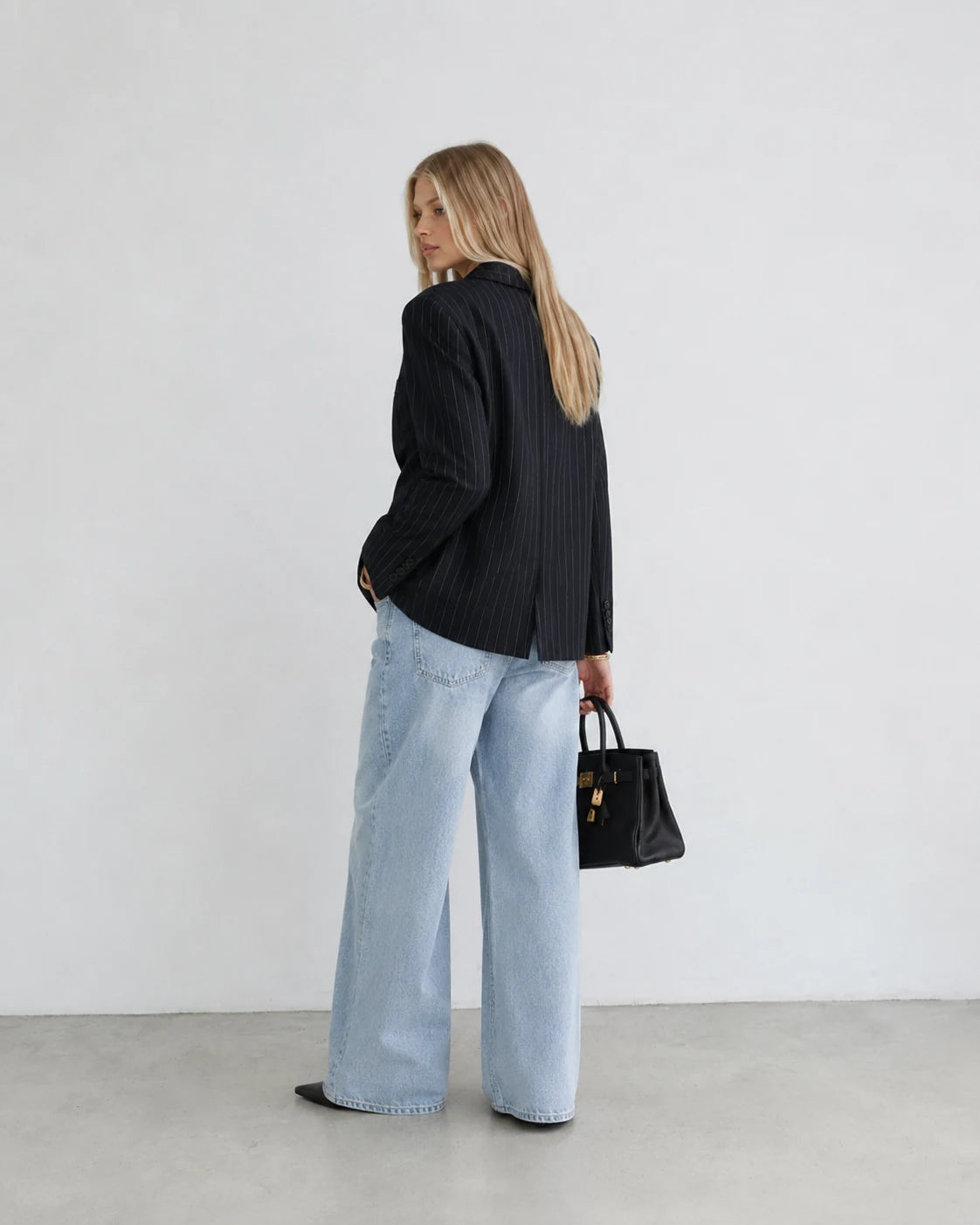 A woman stands in profile wearing a black pinstriped blazer, light-wash wide-leg jeans, and black shoes, holding a black handbag against a plain white wall and gray floor.