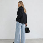 A woman stands in profile wearing a black pinstriped blazer, light-wash wide-leg jeans, and black shoes, holding a black handbag against a plain white wall and gray floor.
