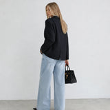 A woman stands in profile wearing a black pinstriped blazer, light-wash wide-leg jeans, and black shoes, holding a black handbag against a plain white wall and gray floor.