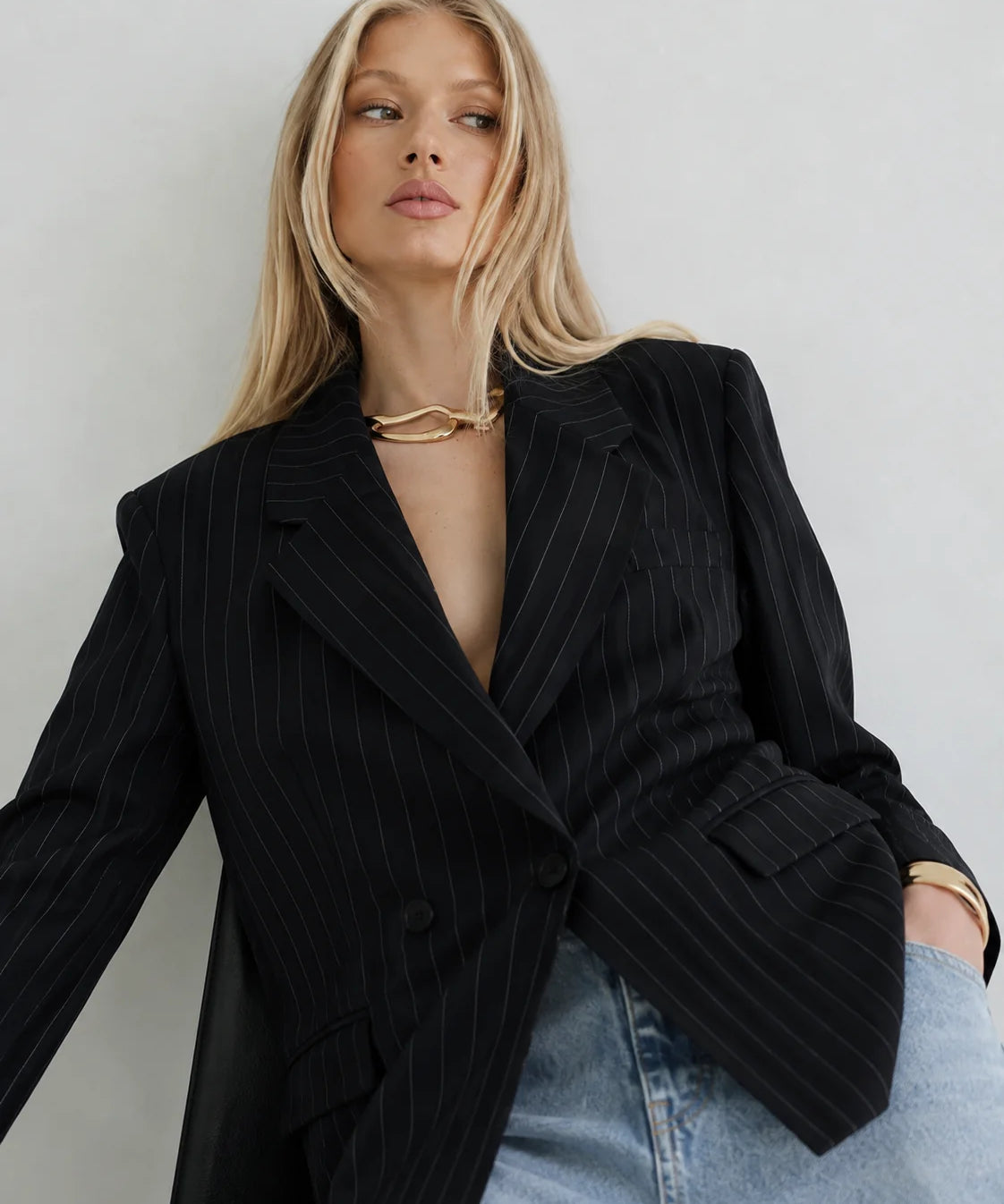 A blonde model wearing a black double-breasted pinstripe blazer over a black dress, styled with light-wash blue jeans and gold jewelry, set against a plain light gray background.