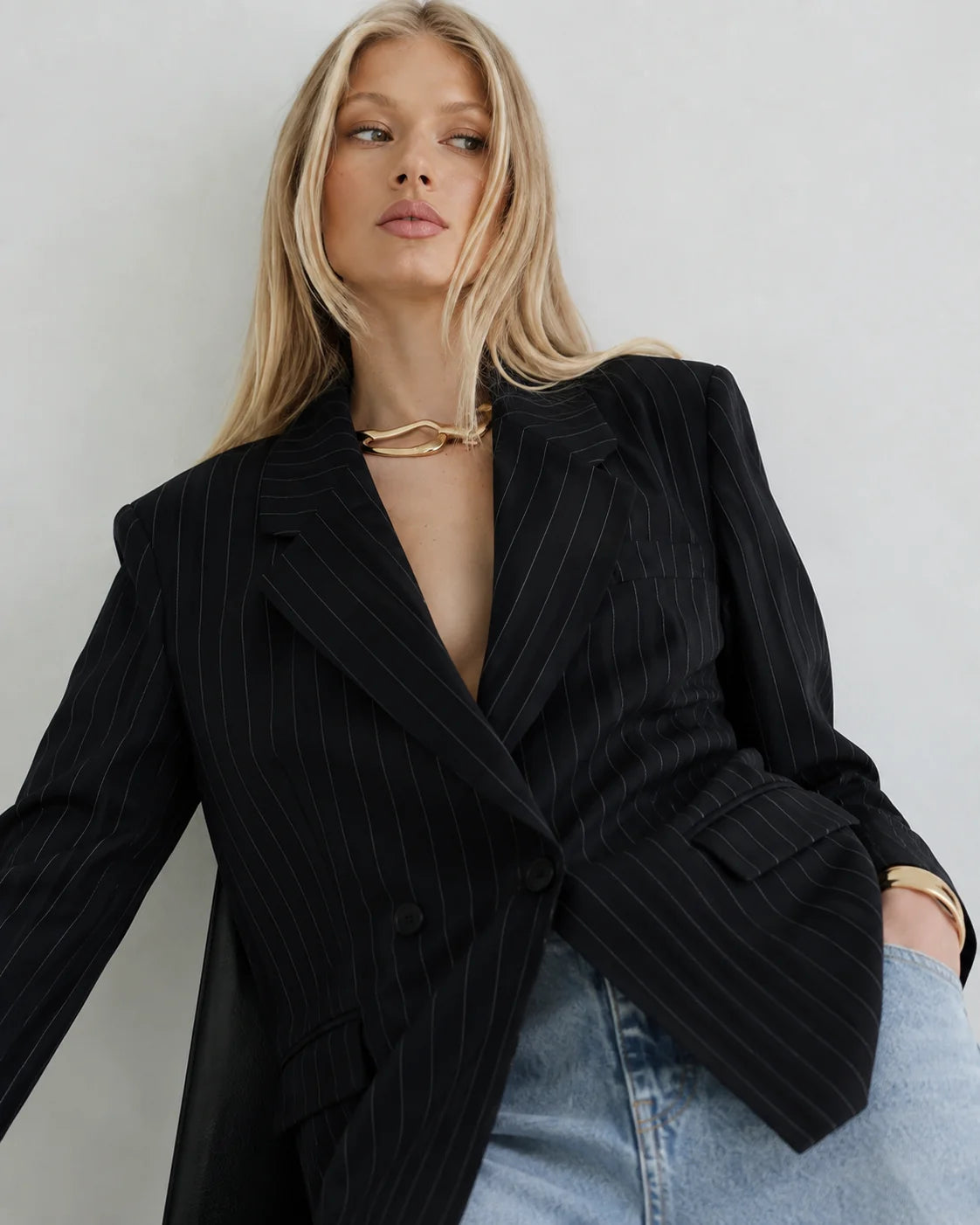 A blonde model wearing a black double-breasted pinstripe blazer over a black dress, styled with light-wash blue jeans and gold jewelry, set against a plain light gray background.