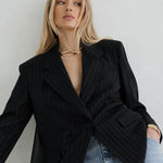 A blonde model wearing a black double-breasted pinstripe blazer over a black dress, styled with light-wash blue jeans and gold jewelry, set against a plain light gray background.