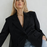 A blonde model wearing a black double-breasted pinstripe blazer over a black dress, styled with light-wash blue jeans and gold jewelry, set against a plain light gray background.