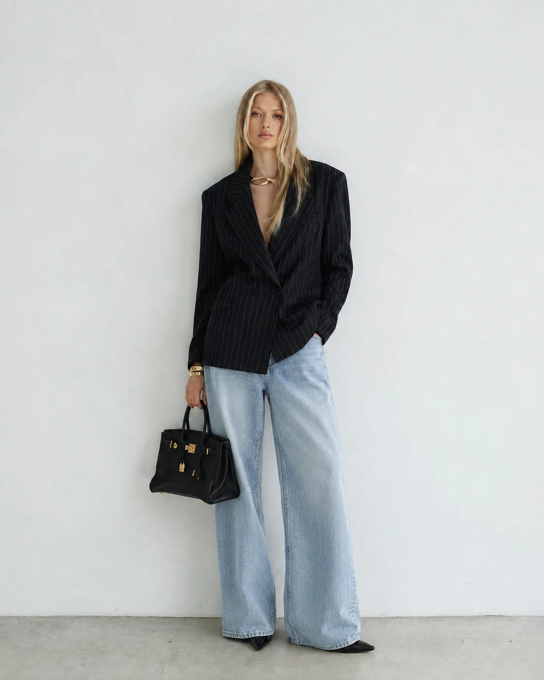 A woman stands against a plain white wall wearing a black pinstriped blazer, wide-leg light blue jeans, black pointed flats, and holding a structured black handbag with gold hardware.