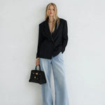 A woman stands against a plain white wall wearing a black pinstriped blazer, wide-leg light blue jeans, black pointed flats, and holding a structured black handbag with gold hardware.