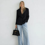A woman stands against a plain white wall wearing a black pinstriped blazer, wide-leg light blue jeans, black pointed flats, and holding a structured black handbag with gold hardware.