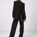 A model from behind wearing a black oversized blazer and matching wide-leg trousers, holding a black clutch bag against a clean white studio background.