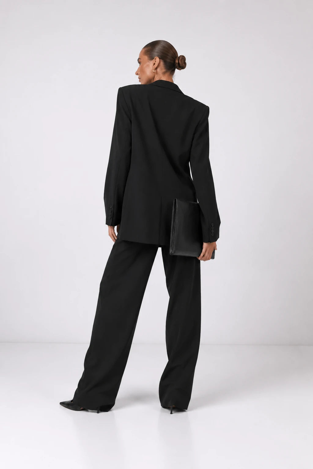 A model from behind wearing a black oversized blazer and matching wide-leg trousers, holding a black clutch bag against a clean white studio background.