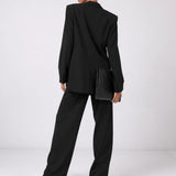 A model from behind wearing a black oversized blazer and matching wide-leg trousers, holding a black clutch bag against a clean white studio background.