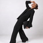 A model in a black tailored suit and black heels poses dynamically on a seamless light gray studio background, holding a black clutch.