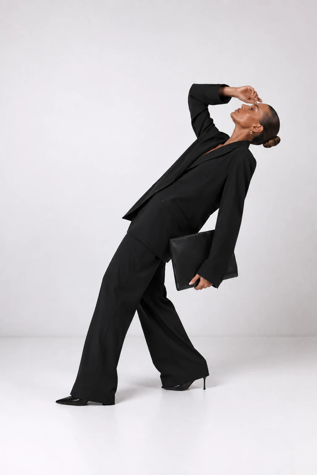 A model in a black tailored suit and black heels poses dynamically on a seamless light gray studio background, holding a black clutch.