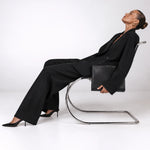 A model in a black blazer and wide-leg trousers sits reclined on a chrome cantilever chair, holding a black leather clutch bag, against a seamless white studio background.