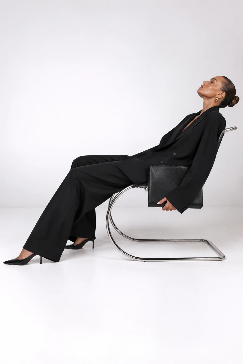 A model in a black blazer and wide-leg trousers sits reclined on a chrome cantilever chair, holding a black leather clutch bag, against a seamless white studio background.