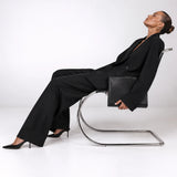 A model in a black blazer and wide-leg trousers sits reclined on a chrome cantilever chair, holding a black leather clutch bag, against a seamless white studio background.