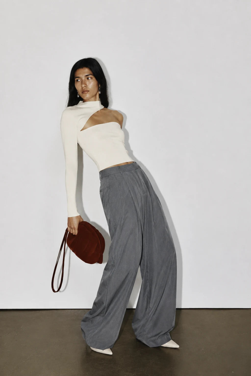 A model stands against a plain white wall wearing a white asymmetrical long-sleeve top, loose gray wide-leg pants, and white pointed shoes, while holding a dark brown shoulder bag.
