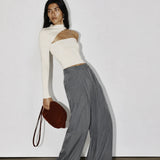 A model stands against a plain white wall wearing a white asymmetrical long-sleeve top, loose gray wide-leg pants, and white pointed shoes, while holding a dark brown shoulder bag.