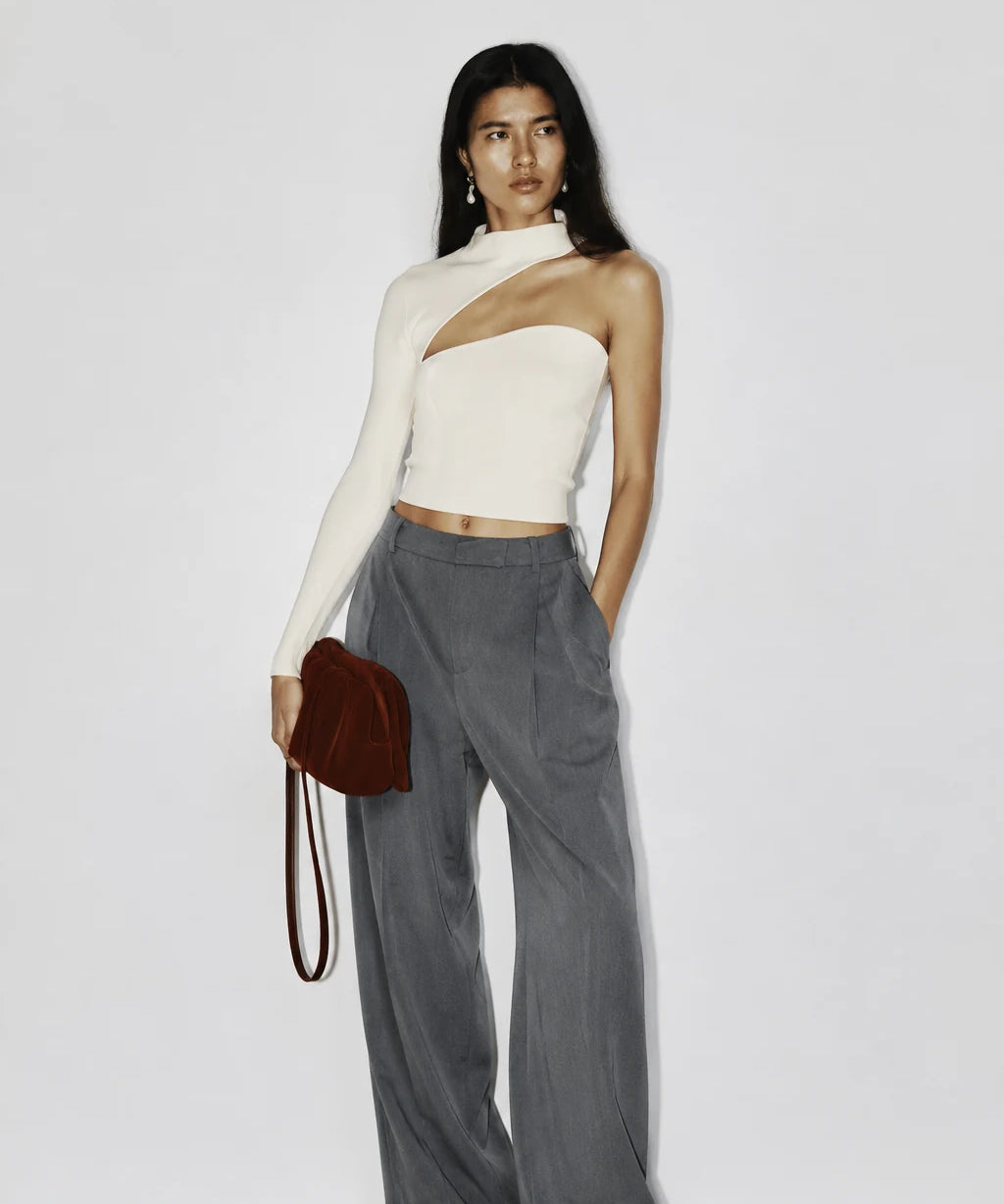 A model stands against a plain light gray wall wearing a one-shoulder cream knit top, high-waisted gray wide-leg trousers, and holding a small dark red shoulder bag.