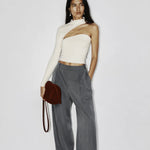 A model stands against a plain light gray wall wearing a one-shoulder cream knit top, high-waisted gray wide-leg trousers, and holding a small dark red shoulder bag.