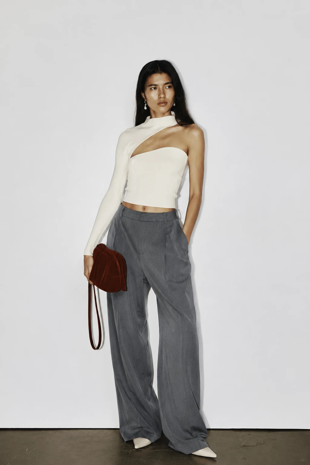 A model stands against a plain light gray wall wearing a one-shoulder cream knit top, high-waisted gray wide-leg trousers, and holding a small dark red shoulder bag.
