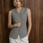 A woman wearing a gray textured sleeveless wrap vest with front buttons and white trousers, standing against a dark wood panel background.