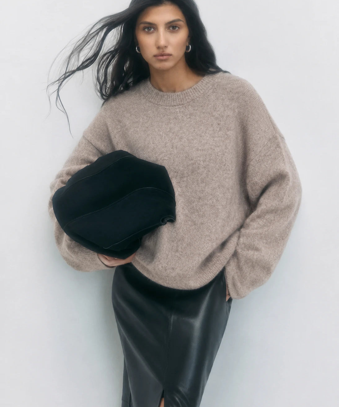 A woman wearing an oversized beige knit sweater and a black leather skirt, holding a black structured handbag against a plain light gray background.