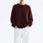 A model wearing a fuzzy, oversized crewneck sweater in deep burgundy, paired with white trousers, against a plain white studio background.
