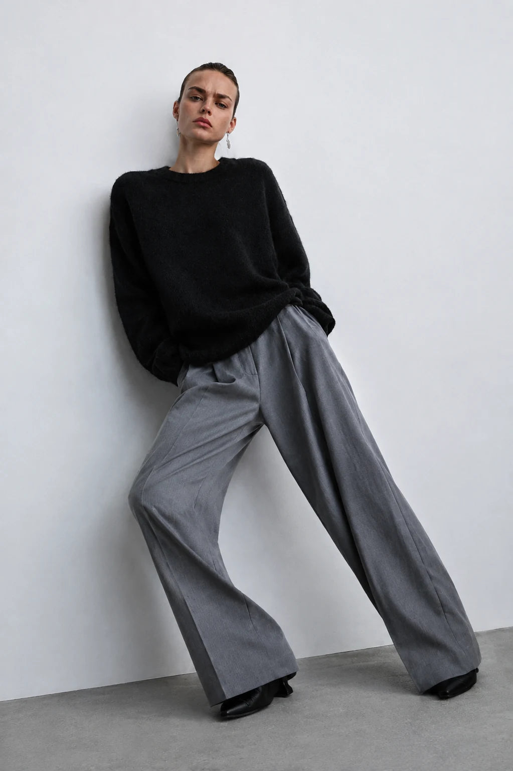 A model wearing a black crewneck knit sweater and wide-leg gray trousers, leaning against a minimalist white wall on a gray floor.