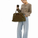 A woman stands against a white studio background holding a black coffee cup and wearing a taupe cropped jacket, wide-leg light blue jeans, and black shoes, while carrying a large olive green suede tote bag.