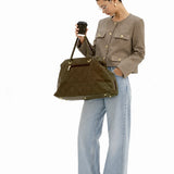 A woman stands against a white studio background holding a black coffee cup and wearing a taupe cropped jacket, wide-leg light blue jeans, and black shoes, while carrying a large olive green suede tote bag.