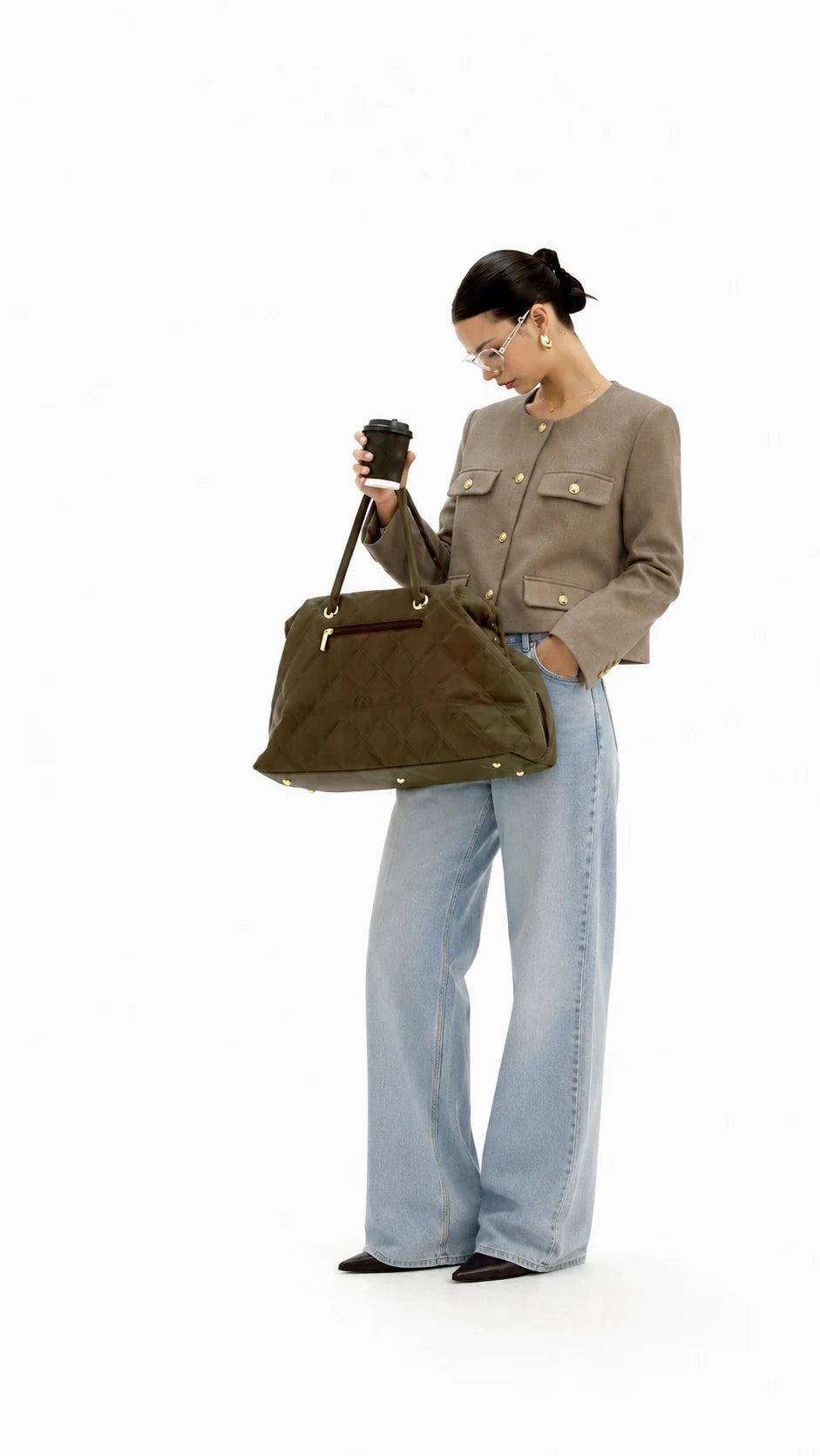 A woman stands against a white studio background holding a black coffee cup and wearing a taupe cropped jacket, wide-leg light blue jeans, and black shoes, while carrying a large olive green suede tote bag.