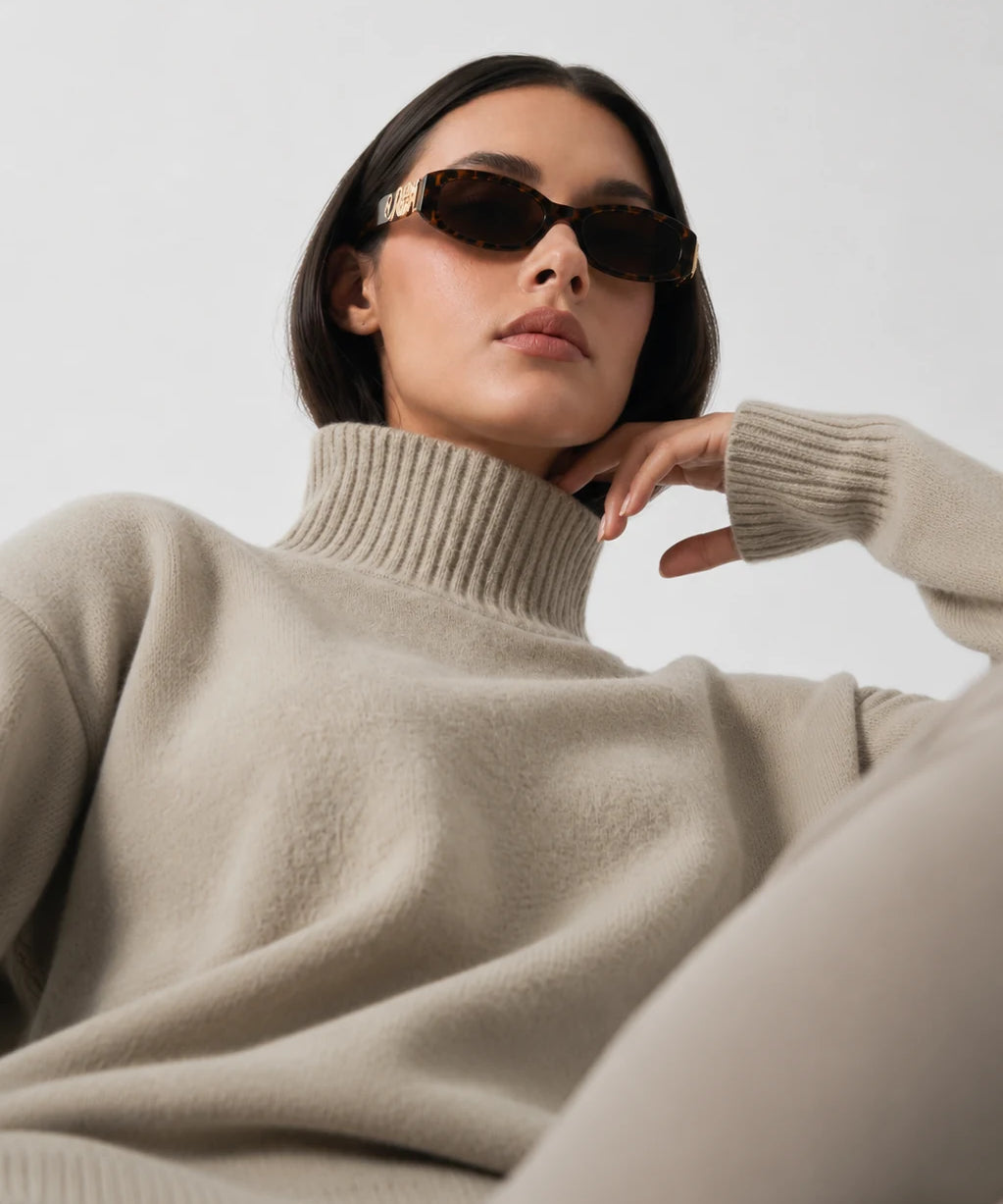 A woman wearing a beige ribbed turtleneck sweater and dark sunglasses, posed against a clean light gray background.