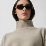 A model with short dark hair is shown from the chest up wearing a light beige ribbed turtleneck sweater and dark brown sunglasses with gold metal frames, set against a plain light gray background.